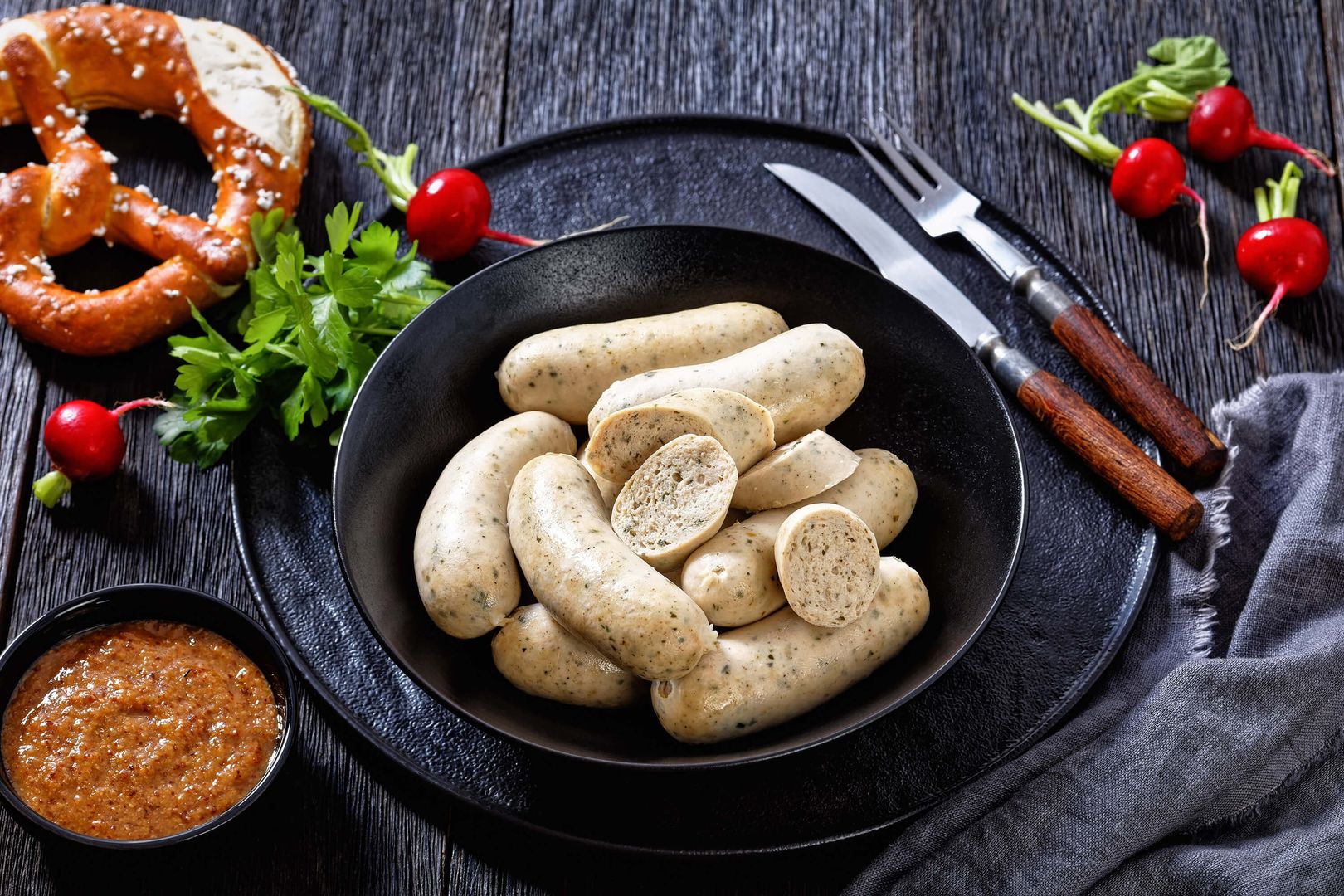 Black plate with white sausages on a black wooden table, next to it is a small bowl of sweet mustard, radishes and a pretzel